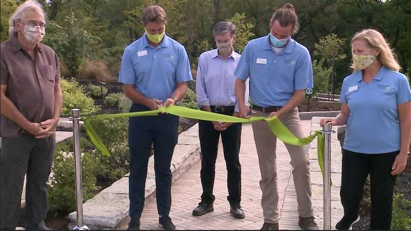 Botanical Garden employees in blue shirts cut a bright green ribbon on a brick pathway.